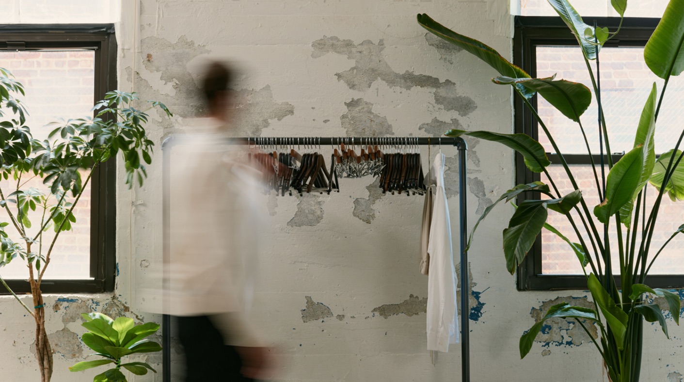 A garment rack of wooden hangers against a weathered plaster wall in a sunlit studio, framed by large plants, with a figure walking past in motion blur